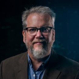 Man with short grey hair with beard wearing glasses, sports coat and denim blue button up shirt