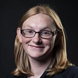 Lady with dark blonde shoulder length hair wearing black top and glasses standing in front of black background